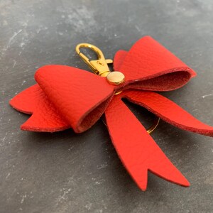 May include: A vibrant red leather bow keyring with a gold-coloured clasp and rivet. The bow is detailed with textured leather and has a classic bow shape. The keyring is a decorative accessory.