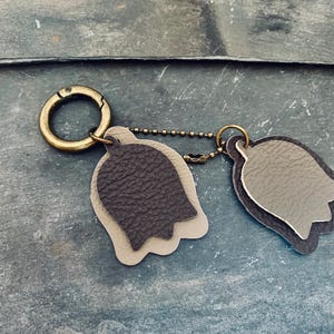 Op de afbeelding: Een leren sleutelhanger met twee tulpvormige bedels in grijstinten en donkergrijs. De bedels zijn bevestigd aan een messingkleurige sluiting en ketting. De sleutelhanger is ontworpen voor sleutels of accessoires.
