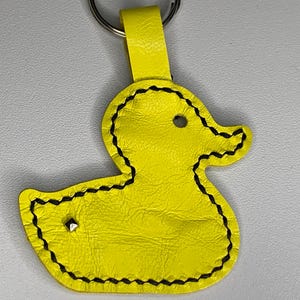 May include: A yellow leather duck keyring with black stitching and a silver rivet. The keyring has a matching yellow strap and a silver ring. The duck has a circular eye and a beak shape.