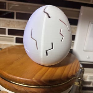 May include: A white, egg-shaped object with jagged cut-out lines, resembling a cracked eggshell. The egg is resting on a wooden lid with a gold-colored metal band. The background features a tiled wall.