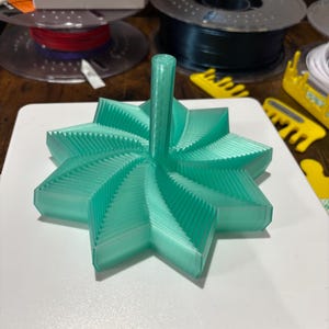 以下が含まれることがあります： 中央の柱が付いた、3Dプリントされたミントグリーンの星型オブジェクト。オブジェクトは、層状の半透明プラスチックでできており、白い表面にあります。デザインは複数のポイントとテクスチャード加工された表面を備えています。