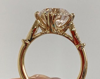 Könnte beinhalten: Ein goldener Verlobungsring mit einem großen, klaren, runden Brillanten, der in einer filigranen, verzierten Fassung sitzt. Die Band ist dünn und hat eine strukturierte Oberfläche.