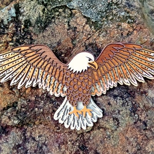 Pu&ograve; includere: Un ritaglio di legno di un'aquila calva con piume marroni e bianche, con le ali spiegate. L'aquila guarda a destra e ha un design dettagliato.