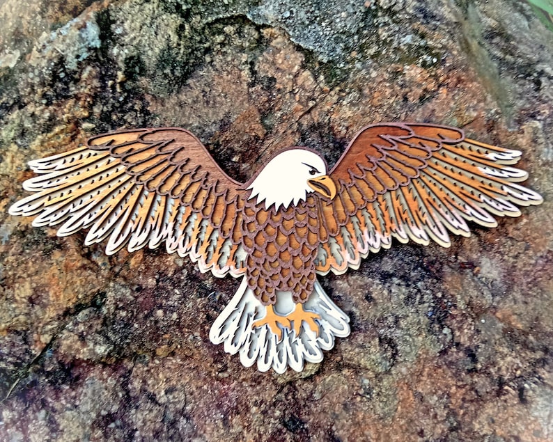 Pu&ograve; includere: Un ritaglio di legno di un'aquila calva con piume marroni e bianche, con le ali spiegate. L'aquila guarda a destra e ha un design dettagliato.