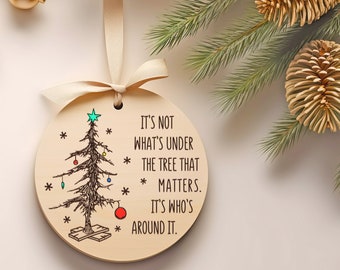 Puede incluir: Un adorno navideño redondo de madera con una ilustración dibujada a mano de un árbol de Navidad con adornos y una estrella roja en la parte superior. El adorno tiene el texto "No es lo que está debajo del árbol lo que importa. Es quién está alrededor."