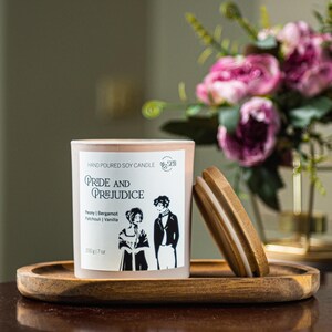 May include: A soy wax candle in a frosted glass jar with a wooden lid, labelled "Pride and Prejudice." The label features a black and white illustration of a couple. The candle sits on a wooden tray, with a bouquet of pink flowers in the background.