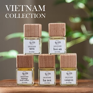 May include: Five small glass bottles with wooden caps, each containing a car diffuser. The bottles are arranged in a pyramid shape. The labels on the bottles read "sen", "hoi an", "vietnamese iced coffee", "ha noi", and "saigon". Each bottle contains 8 ml of liquid.