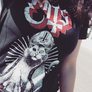 May include: Black t-shirt featuring a graphic of a cat wearing a papal mitre with a cross, surrounded by radiating lines. The word "CAT" is printed in a gothic font at the top. The design is in white and red.