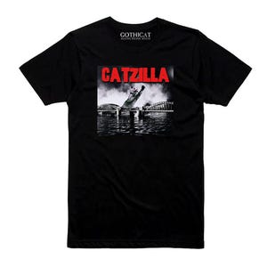 May include: Black t-shirt featuring a graphic of a cat as Godzilla, with the word "CATZILLA" in red. The image shows a cat in a city scene with a bridge and water. The shirt has the text "GOTHICAT" at the top.