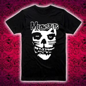 May include: Black t-shirt featuring a white graphic of a skull with cat ears and whiskers. The word "MEOWSFITS" is printed above the skull in a stylized font. The shirt is displayed against a patterned background.