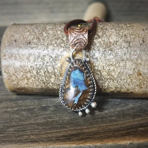 Puede incluir: Un colgante de plata con una piedra de ópalo en forma de lágrima engastada en un engaste de plata. El ópalo tiene un color azul y marrón con una cadena de plata y cobre.