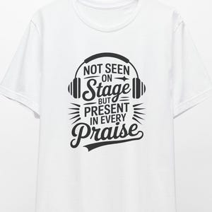 May include: White t-shirt with the text "NOT SEEN ON STAGE BUT PRESENT IN EVERY PRAISE" in black. The design includes headphones and decorative lines. The shirt is hanging on a wooden hanger.