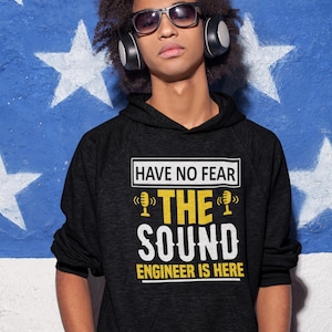 May include: A black hoodie with the text "Have no fear, the sound engineer is here" in yellow. The text is surrounded by a white rectangle. Two yellow microphones are on either side of the word "the".