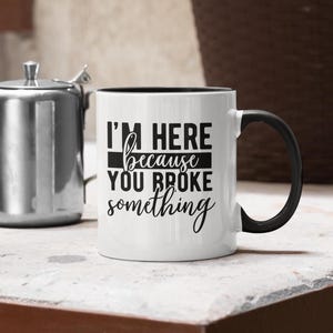 May include: White coffee mug with a black rim and handle. The mug has black text that reads "I'm here because you broke something."