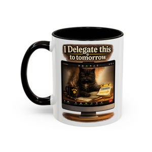 May include: A white ceramic mug with a black handle and interior, featuring a graphic of a computer screen displaying a black cat at a desk with a coffee mug and a calendar. The text above the screen reads I Delegate this to tomorrow.