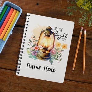 May include: A spiral-bound notebook with a watercolor illustration of a lantern surrounded by flowers. The text "BE THE Light Matthew 5:14" and "Name Here" are printed on the cover. Colored pencils and two pencils are also visible.