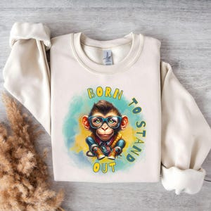 May include: Cream-colored sweatshirt featuring a cartoon monkey wearing glasses and a plaid shirt. The monkey is set against a blue and yellow watercolor background. The text "BORN TO STAND OUT" is printed in green and yellow.