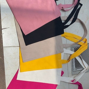 Puede incluir: Un conjunto de cinco bolsas de tela de diferentes colores, incluyendo rosa, negro, beige, amarillo, blanco y fucsia. Las bolsas están hechas de lona y tienen asas largas.