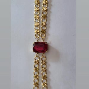 May include: Gold-toned bracelet featuring a rectangular, deep red gemstone at the center. The bracelet has a double-strand chain design with decorative links and a lobster clasp closure. A classic piece of jewelry.