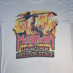 Può includere: Maglietta bianca con una grafica rossa e gialla di un demone che tiene un tridente in fiamme. Il testo "MANOWAR LOCAL CREW HELL ON STAGE" è stampato sotto la grafica.