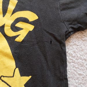 May include: Close-up of a black t-shirt with a yellow graphic design. The design includes the word "ING" and a star. The shirt has a small hole near the sleeve.