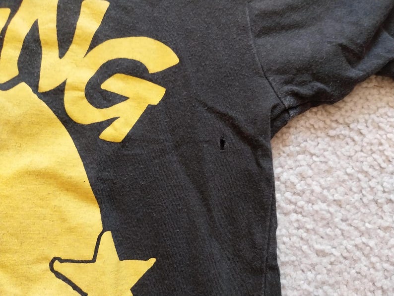 May include: Close-up of a black t-shirt with a yellow graphic design. The design includes the word "ING" and a star. The shirt has a small hole near the sleeve.