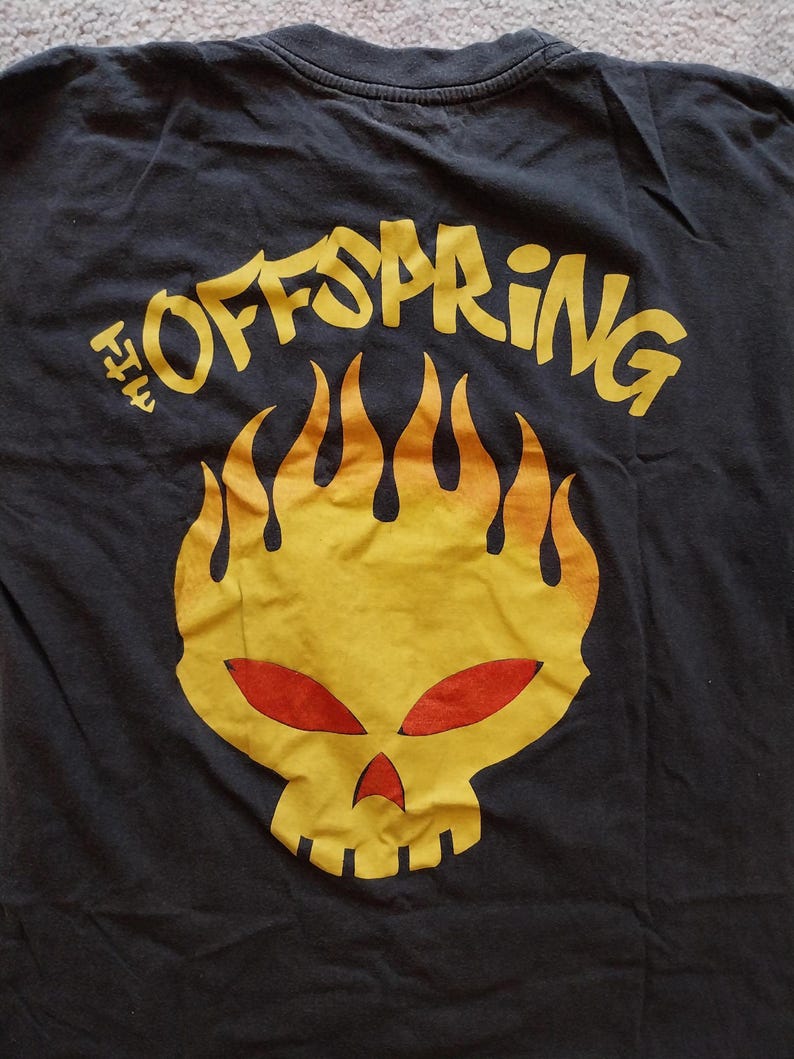 May include: Black t-shirt featuring a yellow and red flaming skull graphic. The text "THE OFFSPRING" is arched above the skull in yellow. The shirt is made of a soft, dark fabric.