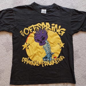 May include: Black t-shirt featuring a graphic of a purple skull with flames, a yellow circle, and the text "The Offspring Original Prankster." Two yellow stars flank the circle. The shirt is short-sleeved.