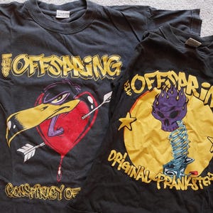 May include: Two black t-shirts featuring The Offspring band graphics. One shirt displays a heart pierced by an arrow with a duck head, the other has a purple skull on a spring. Both shirts have yellow text.