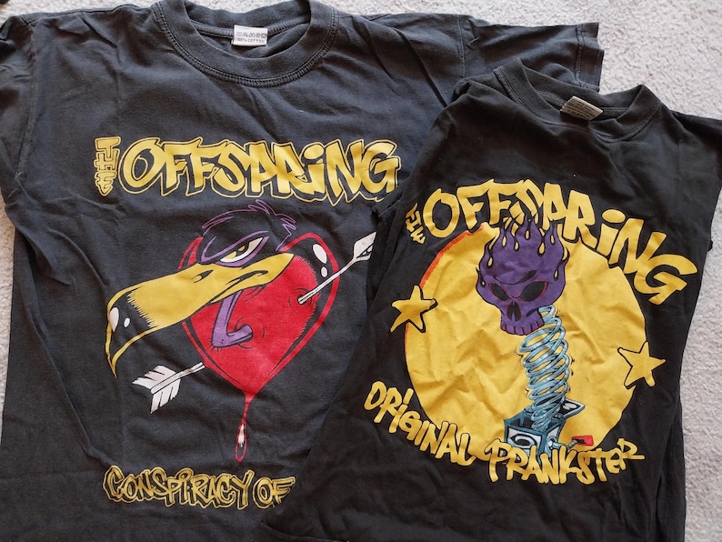 May include: Two black t-shirts featuring The Offspring band graphics. One shirt displays a heart pierced by an arrow with a duck head, the other has a purple skull on a spring. Both shirts have yellow text.