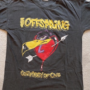 May include: Dark gray t-shirt featuring the band name "The Offspring" in yellow lettering above a graphic of a bird's head piercing a red heart with an arrow. The text "Conspiracy of One" is below the graphic.