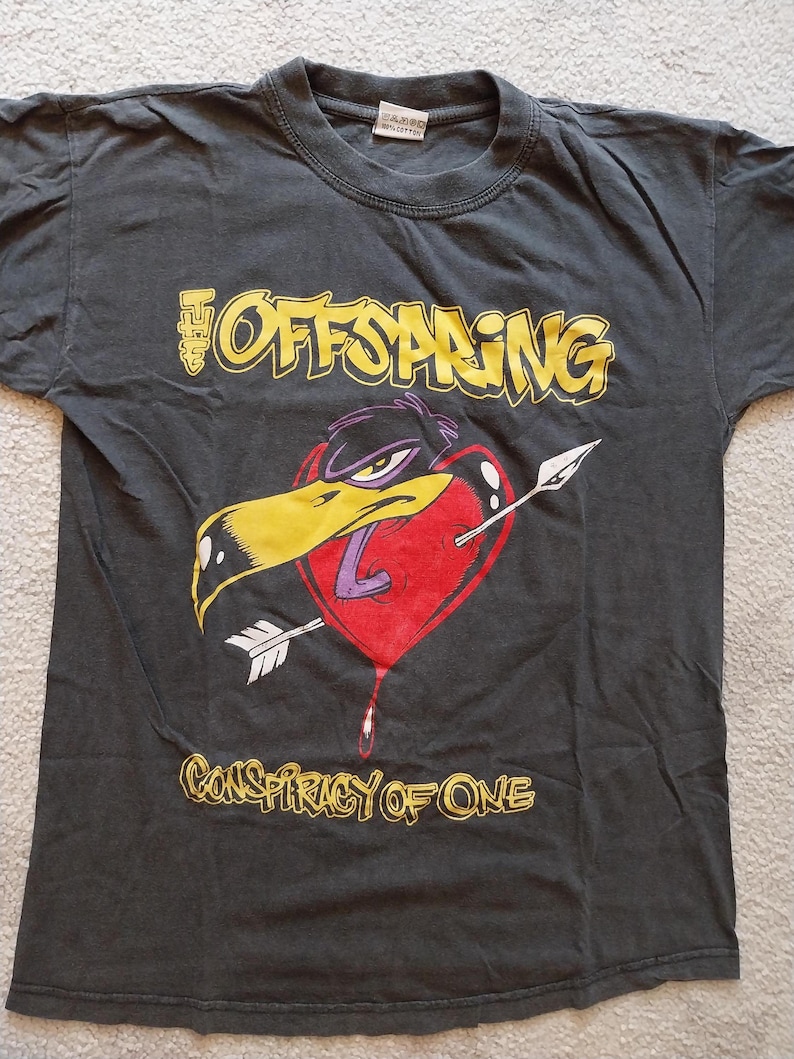 May include: Dark gray t-shirt featuring the band name "The Offspring" in yellow lettering above a graphic of a bird's head piercing a red heart with an arrow. The text "Conspiracy of One" is below the graphic.