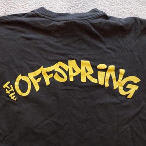 May include: Black t-shirt with the word "OFFSPRING" in yellow, arched across the back. The band's name is in a stylized, graffiti-like font. The shirt is made of a soft material.