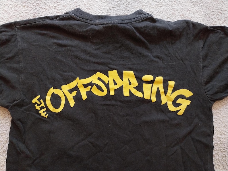 May include: Black t-shirt with the word "OFFSPRING" in yellow, arched across the back. The band's name is in a stylized, graffiti-like font. The shirt is made of a soft material.