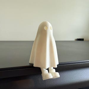 May include: A white ghost figurine with a draped sheet design, featuring two eye holes and small feet. The ghost is sitting on a dark surface, creating a whimsical and spooky decorative item.