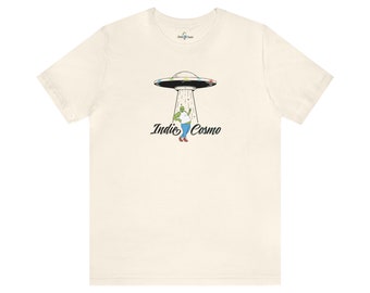 以下が含まれることがあります： 緑色の宇宙人が空飛ぶ円盤の下で踊っている白いTシャツ。宇宙人の下に「Indie Cosmo」という文字が印刷されています。