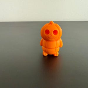 以下が含まれることがあります： 笑顔と腕を持つカボチャの形をした、小さなオレンジ色のハロウィーンをテーマにした置物。この置物には、丸い目の穴と笑顔の口がある彫刻された顔があります。体は垂直線で区切られています。