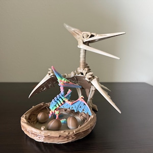 May include: A detailed sculpture of a Pterodactyl skeleton with a colorful baby Pterodactyl in a nest with eggs. The sculpture is beige and brown, with the baby Pterodactyl featuring rainbow colors. The nest is brown.