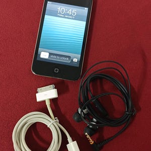 May include: Black iPod touch with a screen displaying the time 10:45 and the date Friday, January 2. Includes a white charging cable, black earbuds, and a red background.