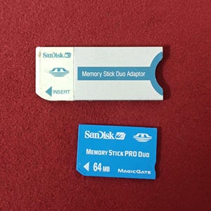 Op de afbeelding: Twee Sandisk geheugenkaarten op een rode ondergrond. Eén is een witte Memory Stick Duo Adapter met blauwe accenten. De andere is een blauwe Memory Stick Pro Duo met 64 MB capaciteit en MagicGate.