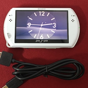 Puede incluir: Consola de juegos portátil Sony PSP blanca con pantalla de reloj. La pantalla muestra una esfera de reloj con la hora a la 1:01. Un cable de carga USB negro está enrollado debajo de la consola. El logotipo de PSP es visible en la parte inferior.