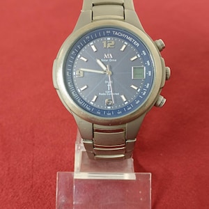 May include: A silver wristwatch with a blue face, displaying the text "MA Solar Drive" and "TACHYMETER". The watch has a metal strap and is presented on a clear stand against a red backdrop. The watch has a digital display.