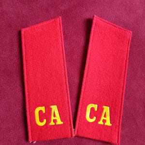 Może przedstawiać: Dwa czerwone naramienniki z żółtymi haftowanymi literami "CA". Prostokątne deski wykonane są z materiału przypominającego filc i są obszyte na krawędziach. Tło jest w kolorze głębokiej czerwieni.