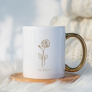 May include: White ceramic mug with a gold handle. The mug has a gold outline of a rose and the date 06.2024 printed on the front.