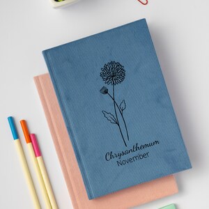 May include: Two notebooks stacked on top of each other. The top notebook is blue with a black line drawing of a chrysanthemum flower and the text "Chrysanthemum November". The bottom notebook is pink.