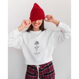May include: White cropped sweatshirt with a black line drawing of a water lily and the text "Water Lily July". The model is wearing a red beanie and red and black plaid pants.