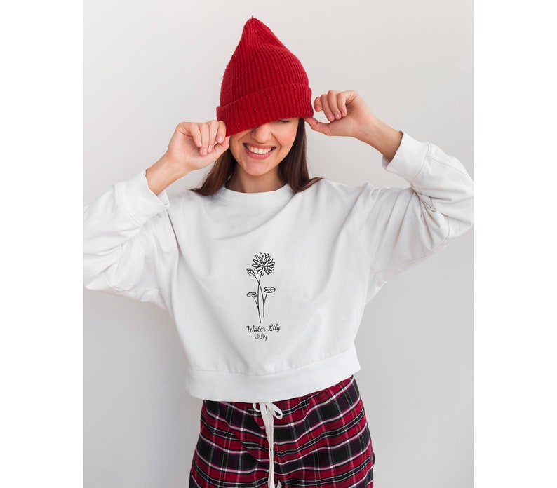 May include: White cropped sweatshirt with a black line drawing of a water lily and the text "Water Lily July". The model is wearing a red beanie and red and black plaid pants.