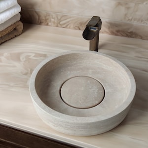 May include: A round, light beige stone bathroom sink with a single faucet. The sink is set on a countertop with a light brown wood grain pattern.