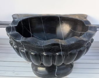 May include: A large, black marble planter with white veining. The planter has a scalloped rim and a pedestal base. The exterior is carved with vertical, rounded flutes. The interior is a deep bowl shape.