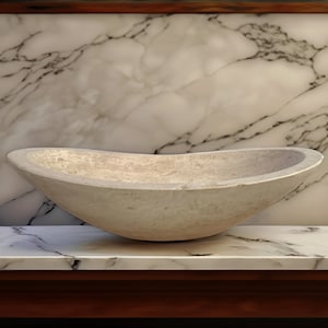 May include: A large, oval-shaped, light beige stone sink. The sink is sitting on a white marble countertop.